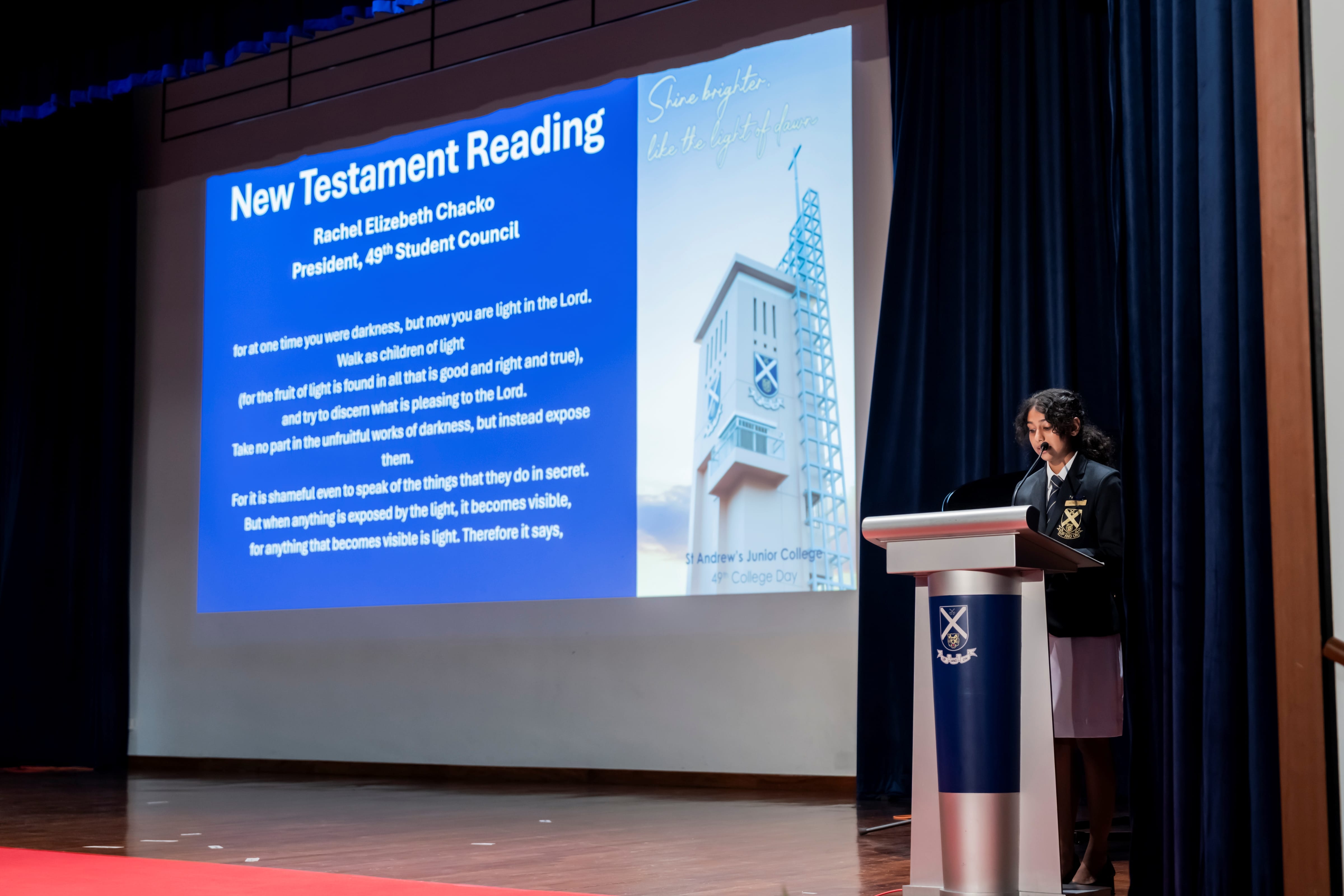 President of the 49th Student Council, Rachel Elizabeth Chacko, stands at a podium delivering a scripture reading next to a projection screen displaying the text and her name.
